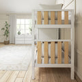 Bright neutral kids bedroom with white wooden bunk bed featuring natural wood slats, white dresser under large window, table lamp and potted plant
