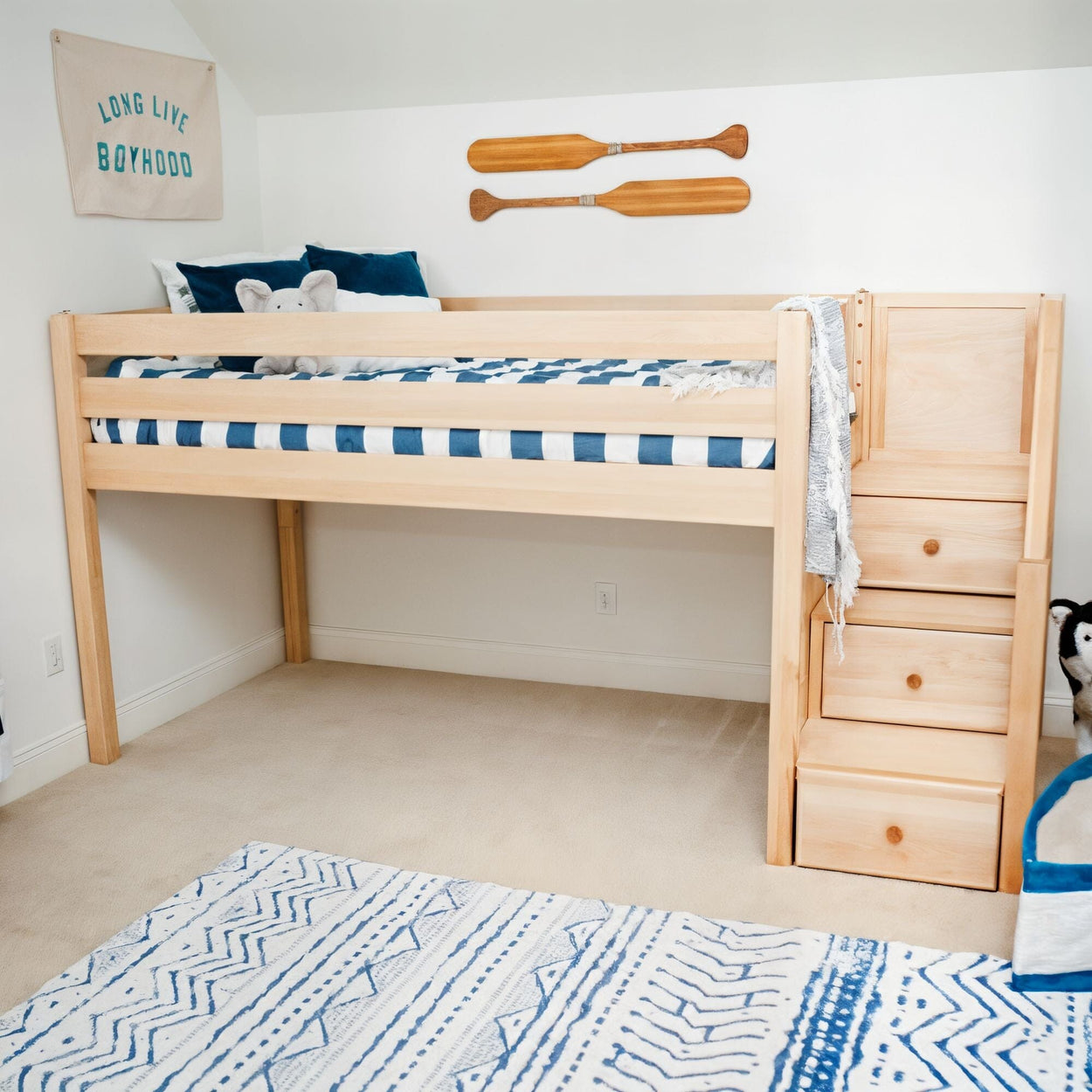 Light wood kids' loft bed with built-in storage stairs, navy striped bedding, wooden paddle wall decor and blue patterned rug