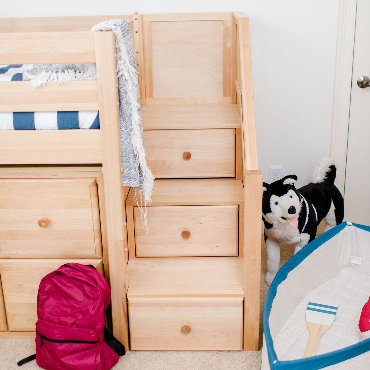 Wooden loft bunk bed with stairs and built-in drawers in a bright kids' bedroom, navy-striped bedding, pink backpack and stuffed husky toy