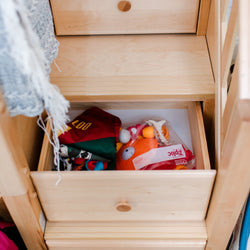 Light pine dresser drawer open showing folded children's clothes, colorful plush toys and a sealed storage bag in a tidy kids' bedroom