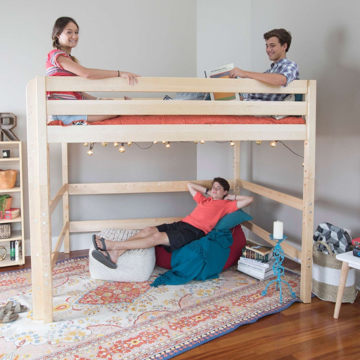 Wooden loft bed in teen bedroom with two teens on top and one lounging below on a beanbag; cozy space-saving design with string lights