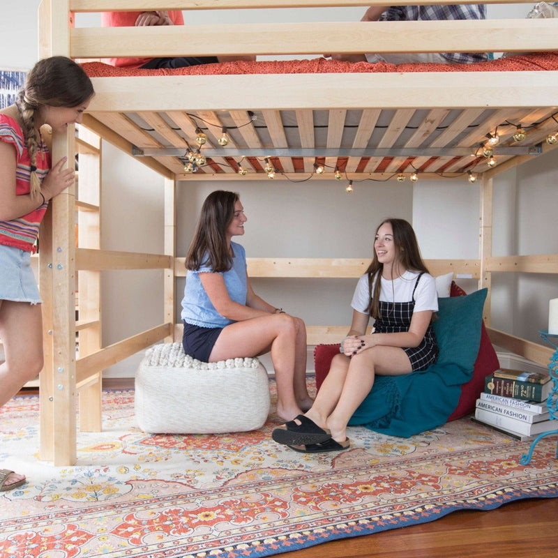 Teen girls in a dorm room gathered under a wooden loft bed with string lights, cozy seating, pouf, patterned rug and stacked books — college dorm decor