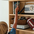 Child reaching for a book on a light wood bookshelf with children's books, vintage radio, wicker basket, Xbox console and football