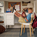 Smiling young boy holding a football at a desk in a study nook with corkboard, lamp, backpack and sports wall art