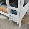 Close-up of a white wooden bunk bed ladder and slatted rail with a blue chunky knit blanket and tan bedding on the lower mattress, set on hardwood floor with a patterned area rug