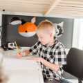Elementary-age boy studying at a bright modern desk, writing with a pencil while using a tablet for digital learning and homework