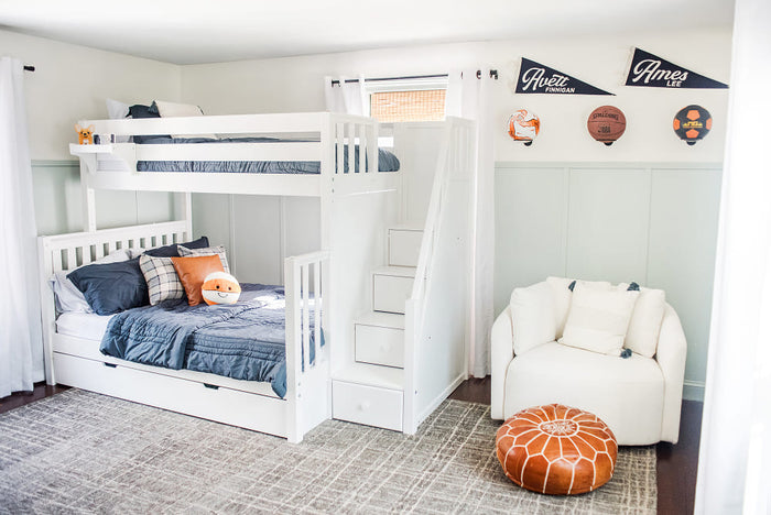 Bright kids' bedroom with modern white bunk bed and stairs, navy bedding, trundle, armchair, pouf and sports pennants.