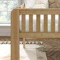 Light wood baby crib with vertical slats, neutral striped bedding, textured throw pillows, tassel rug and nursery window
