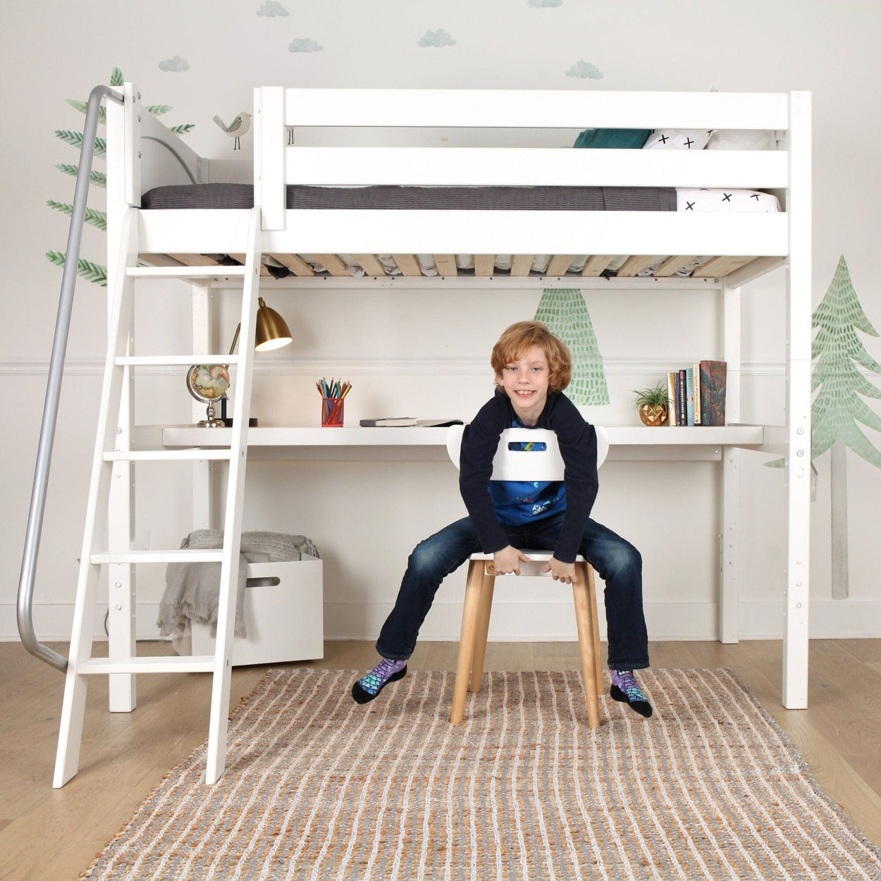 White kids loft bed with desk and ladder, built-in study area under bed with lamp, books and globe, child sitting on stool in children's bedroom