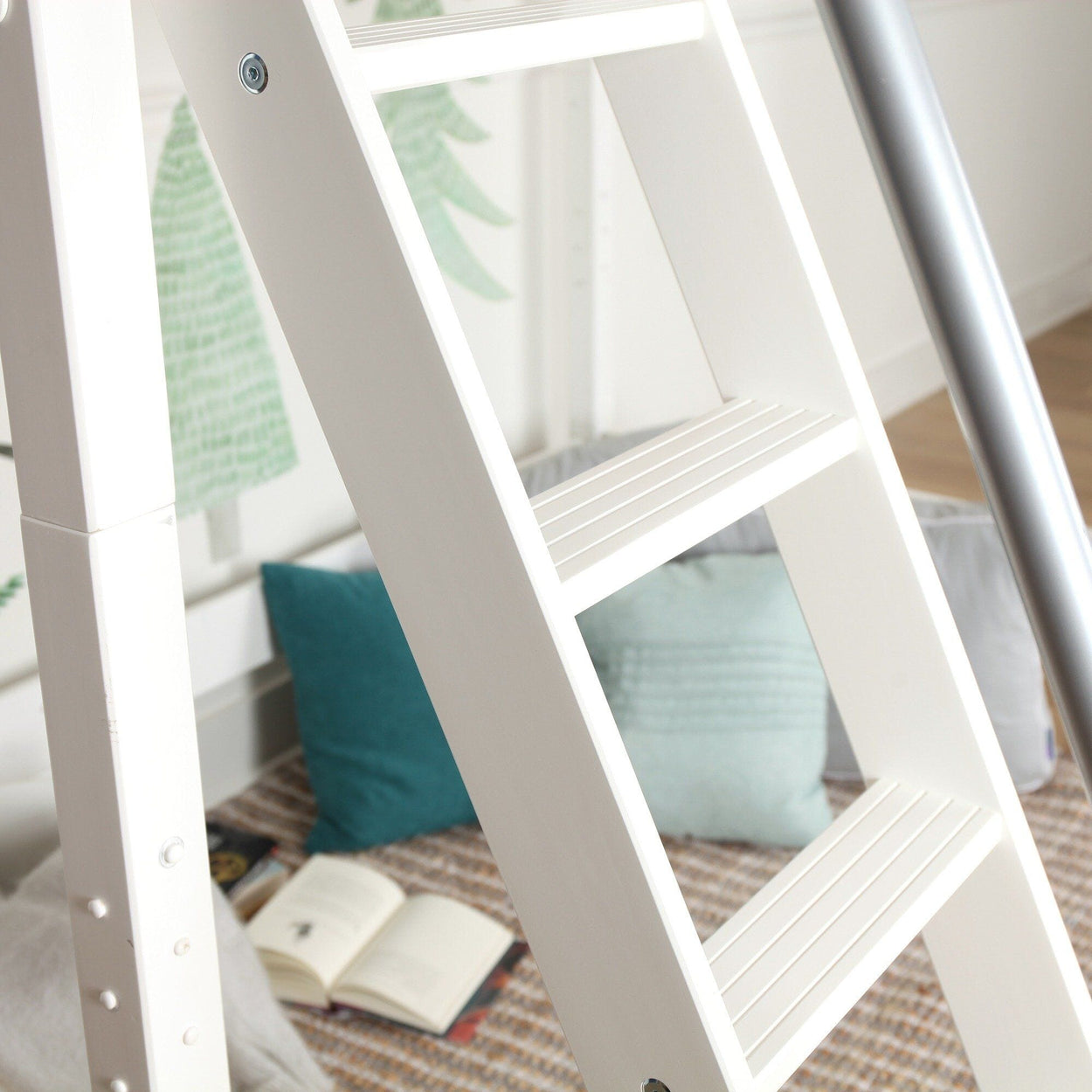 White wooden ladder in a children's bedroom reading nook with teal pillows, open book on woven rug and green tree wall mural