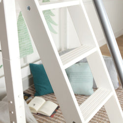 White wooden ladder in a children's bedroom reading nook with teal pillows, open book on woven rug and green tree wall mural