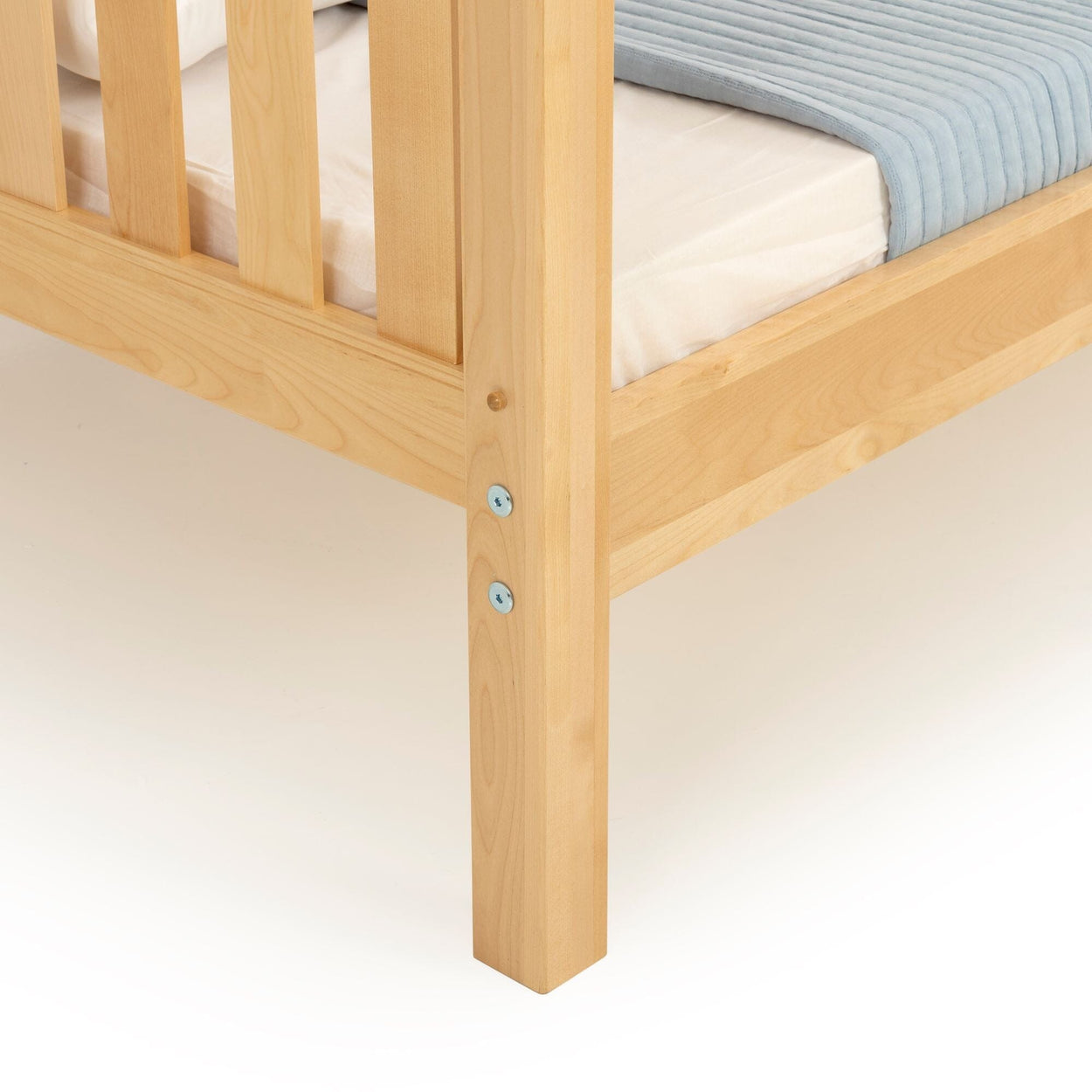 Close-up of light natural solid wood bed frame corner with slatted side rail, sturdy leg and visible metal fasteners, mattress with light blue quilt