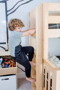Curly-haired young boy climbing a natural wood bunk bed ladder in a bright modern kids' bedroom
