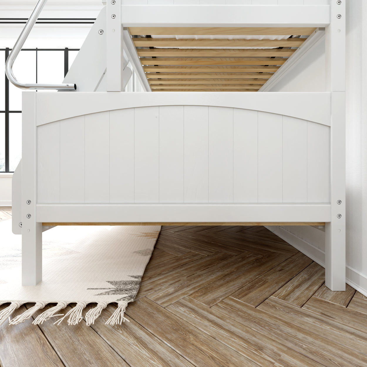 White wooden bunk bed frame with natural wood slats, placed on a chevron-patterned hardwood floor next to a cream-colored fringed rug in a bright room.