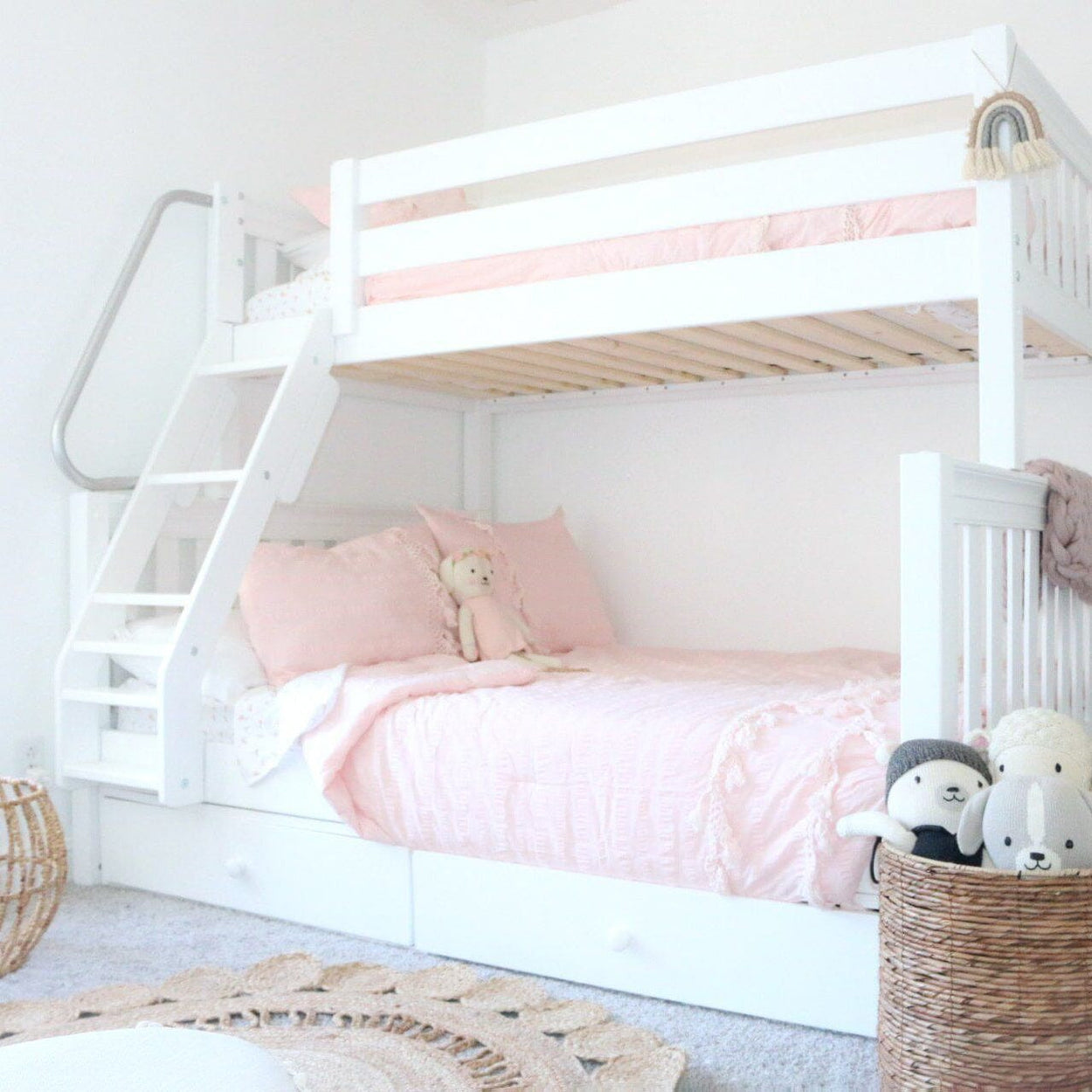 White wooden bunk bed with pink ruffled bedding, under-bed storage drawers, wicker toy basket and plush animals in a cozy kids' bedroom