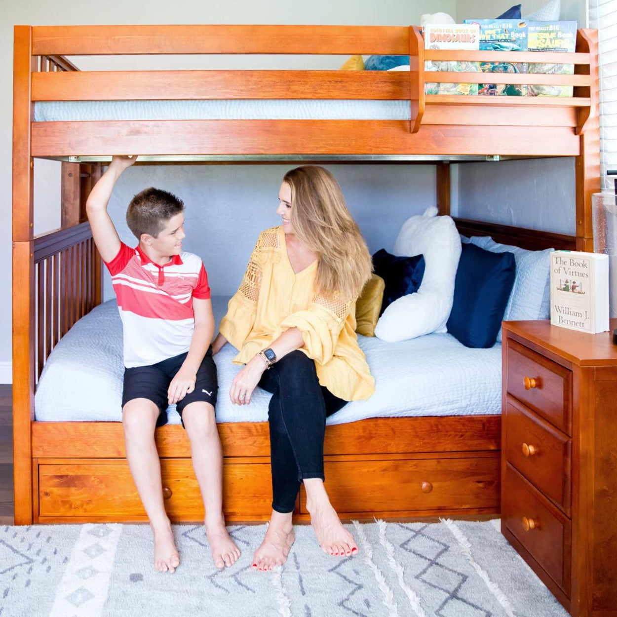 Mother and son sitting on a wooden bunk bed with books and decor in a cozy bedroom setting.