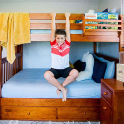 Child hanging from wooden bunk bed ladder in colorful bedroom with books and plush toys.