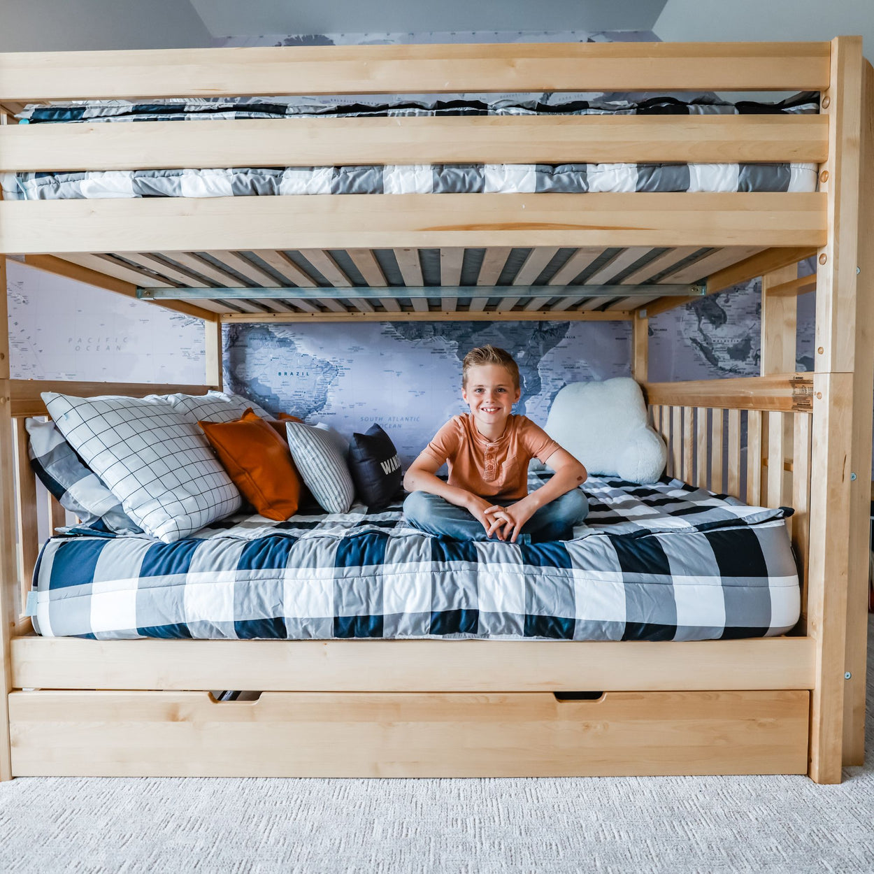 Child sitting on a stylish wooden bunk bed with a world map mural backdrop and decorative pillows.