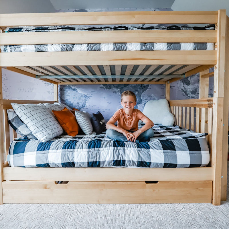 Smiling boy sitting on a wooden twin-over-twin bunk bed with gray plaid bedding, decorative pillows, under-bed storage drawers and world map wall mural in a kids' bedroom