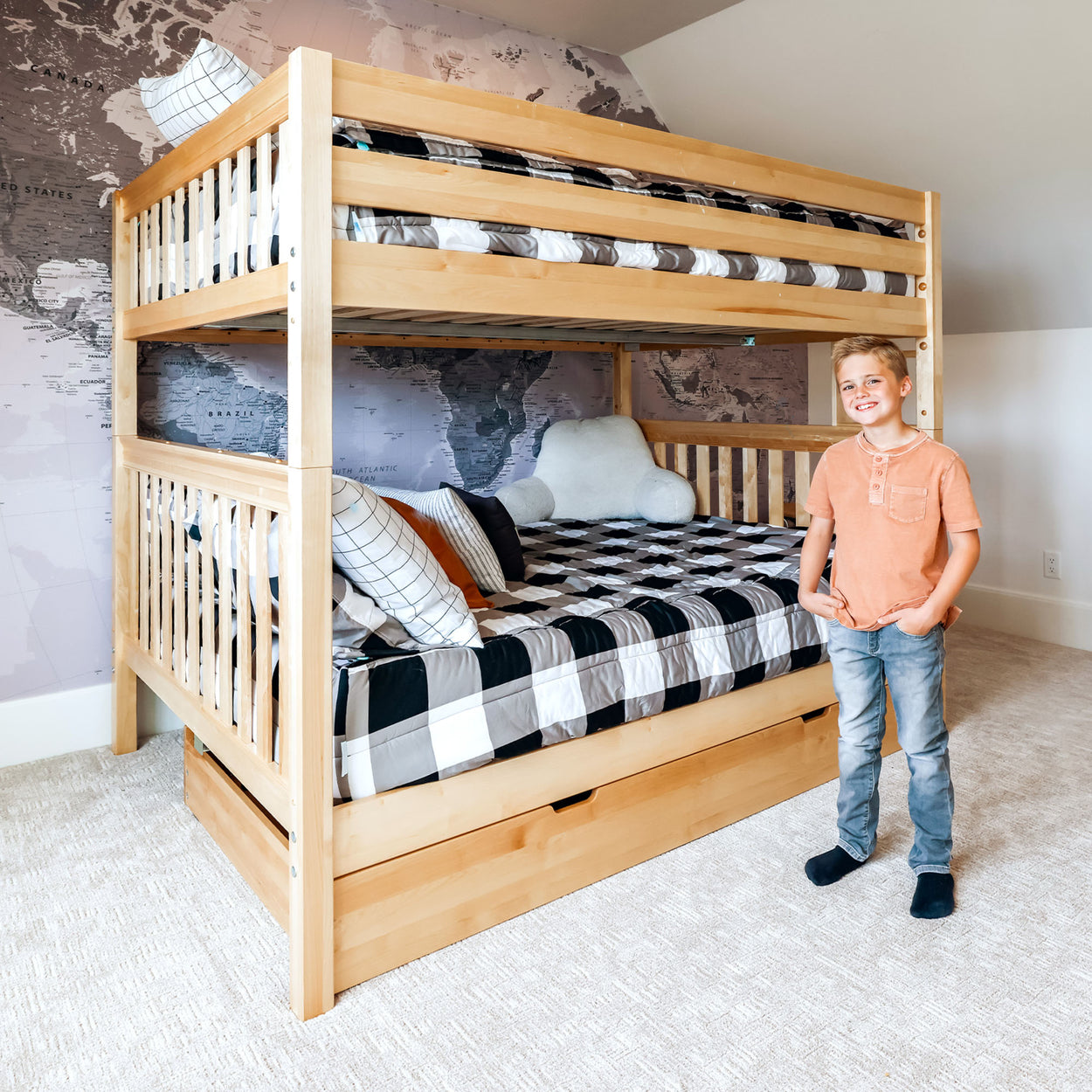 Boy standing next to a wooden bunk bed with checkered bedding in a modern bedroom with a world map wallpaper.