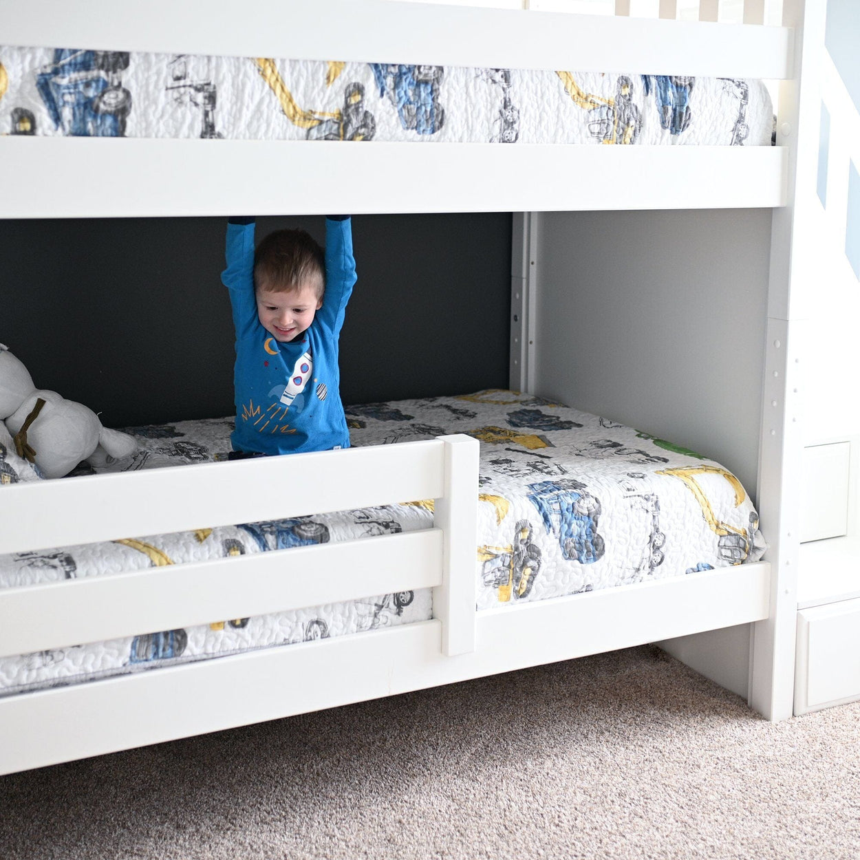 Young child in blue rocket pajamas hanging from the top bunk of a white wooden bunk bed with construction-truck patterned quilt in a bright modern kids' bedroom