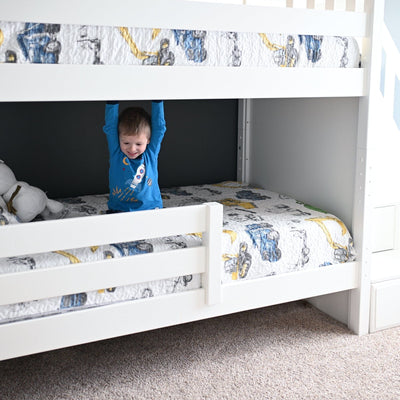 Young child in blue rocket pajamas hanging from the top bunk of a white wooden bunk bed with construction-truck patterned quilt in a bright modern kids' bedroom