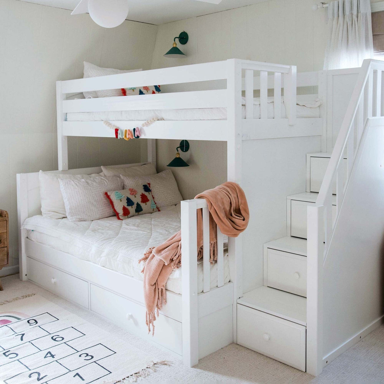 White wooden bunk bed with staircase storage drawers in bright kids' bedroom, cozy pink throw, decorative pillows and hopscotch rug, modern children's room decor