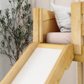Wooden indoor kids' bed slide in natural pine with soft pink mattress and white pillow next to a leafy indoor plant, modern neutral nursery decor