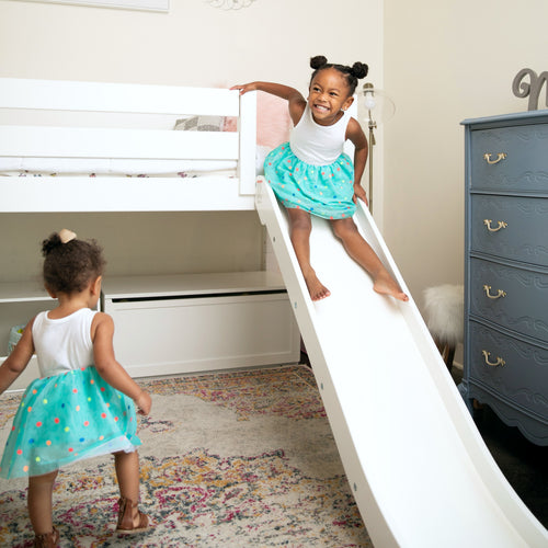 Smiling toddler girl sliding down a white bunk bed slide in a bright kids bedroom while another child plays on a colorful rug — bunk bed with slide, children playing, playroom decor