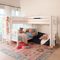 Two children playing on a white kids bunk bed in a cozy girls' bedroom with pastel bedding, plush toys, string lights, and a patterned rug
