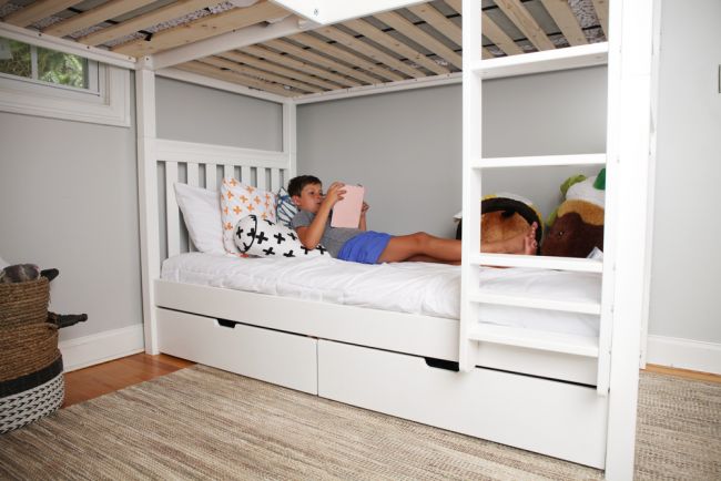 Boy reading a tablet on a white lower bunk bed with pull-out storage drawers in a modern gray kids' bedroom