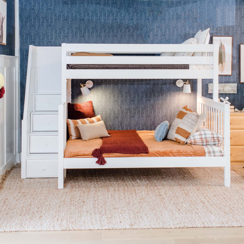 White wooden kids bunk bed with built-in storage stairs in a children's bedroom, rust and beige bedding, throw pillows and wall lamps against blue patterned wallpaper