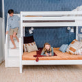 Modern kids' bedroom with white bunk bed and built-in storage stairs, navy patterned wallpaper, orange quilted bedding, decorative pillows and wall-mounted reading lights