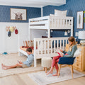 Cozy kids' bedroom with white wooden bunk bed, blue chevron wallpaper, knit pouf and textured rugs, two children reading and using a mobile device
