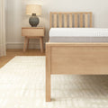 Modern minimalist bedroom with wooden bed frame, white mattress, matching nightstand, lamp, and light-colored rug.