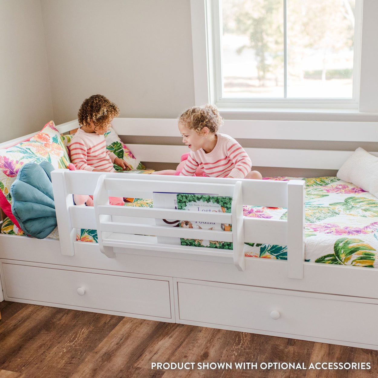 Children playing on a white toddler bed with safety rail and floral bedding in bright bedroom.
