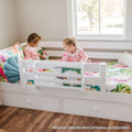 White toddler bed with safety rail and built-in storage drawers in a bright kids' bedroom, two children reading on colorful floral bedding by a window