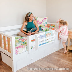 Convertible white toddler bed with storage drawers and built-in book organizer, mother reading to toddler on colorful tropical bedding in bright nursery
