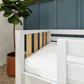 Close-up of a modern nursery with a white toddler bed featuring wood slats and white mattress, teal paneled accent wall, and fiddle leaf fig in a terracotta pot