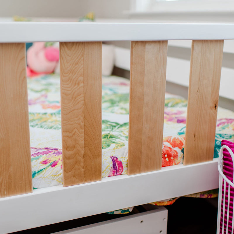 Close-up of a modern white-and-natural-wood baby crib railing with vertical slats and a colorful floral baby quilt in a bright nursery