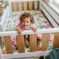 Smiling toddler peeking over white wooden crib railing in sunlit nursery with colorful floral bedding and pink striped sweater