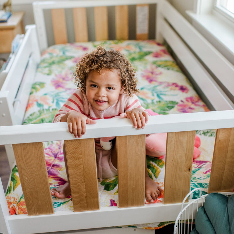 Smiling toddler peeking over white wooden crib railing in sunlit nursery with colorful floral bedding and pink striped sweater