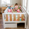 Two children reading on a white wooden toddler bed with a colorful floral quilt and pink plush toys in a cozy nursery