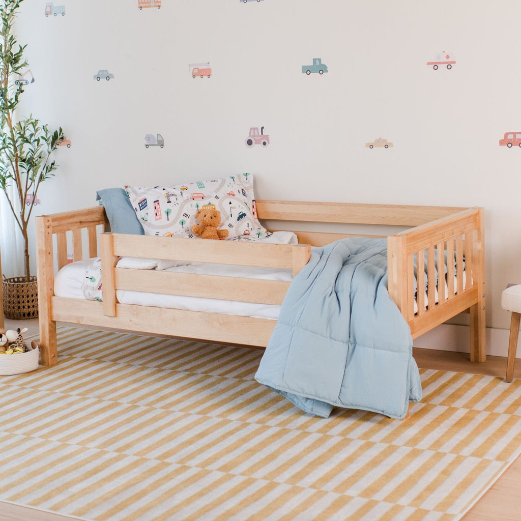 Wooden toddler bed in a nursery with blue comforter, teddy bear, patterned pillow, playful car wall decals and yellow striped area rug