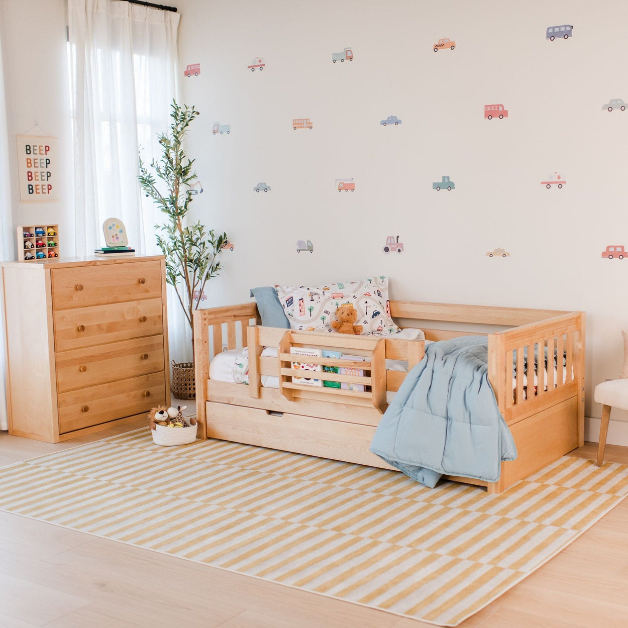 Wooden toddler bed with safety rail and under-bed storage in a bright kids bedroom, styled with a blue comforter and plush toys, matching wooden dresser, car wall decals, striped yellow rug and potted plant for modern nursery decor